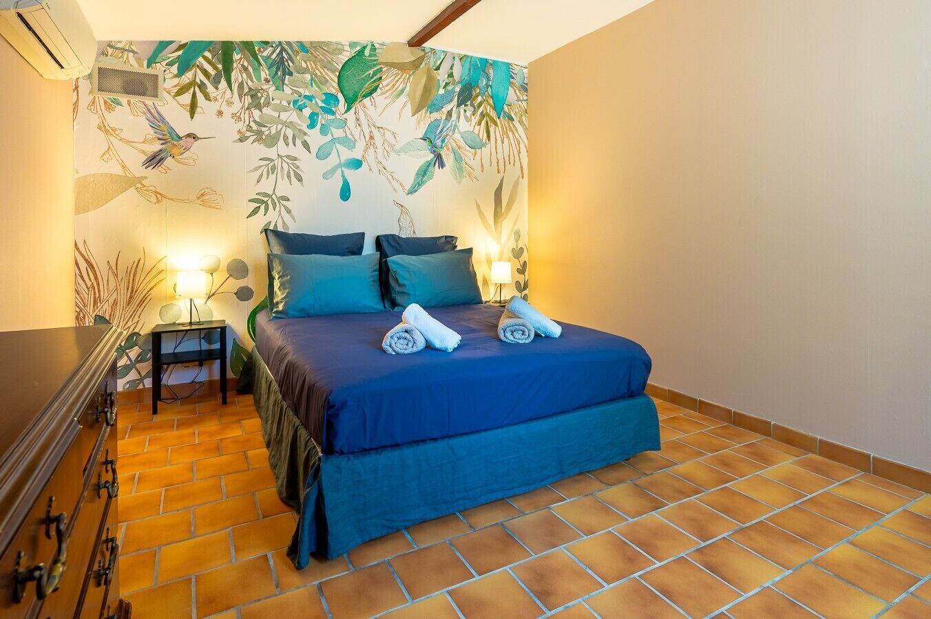 Ein Schlafzimmer mit einem Bett, Kissen, einem Nachttisch, einer Lampe und einer Wand mit einem Blattmuster.