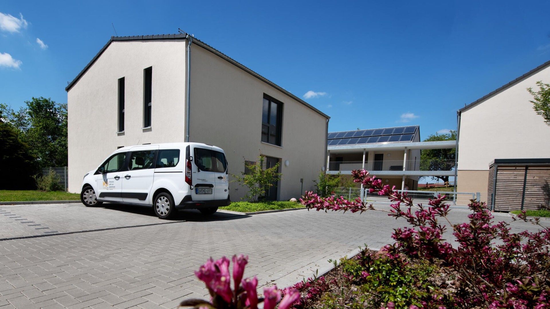 A modern building with a white van parked in front, solar panels on the roof, and a paved driveway.