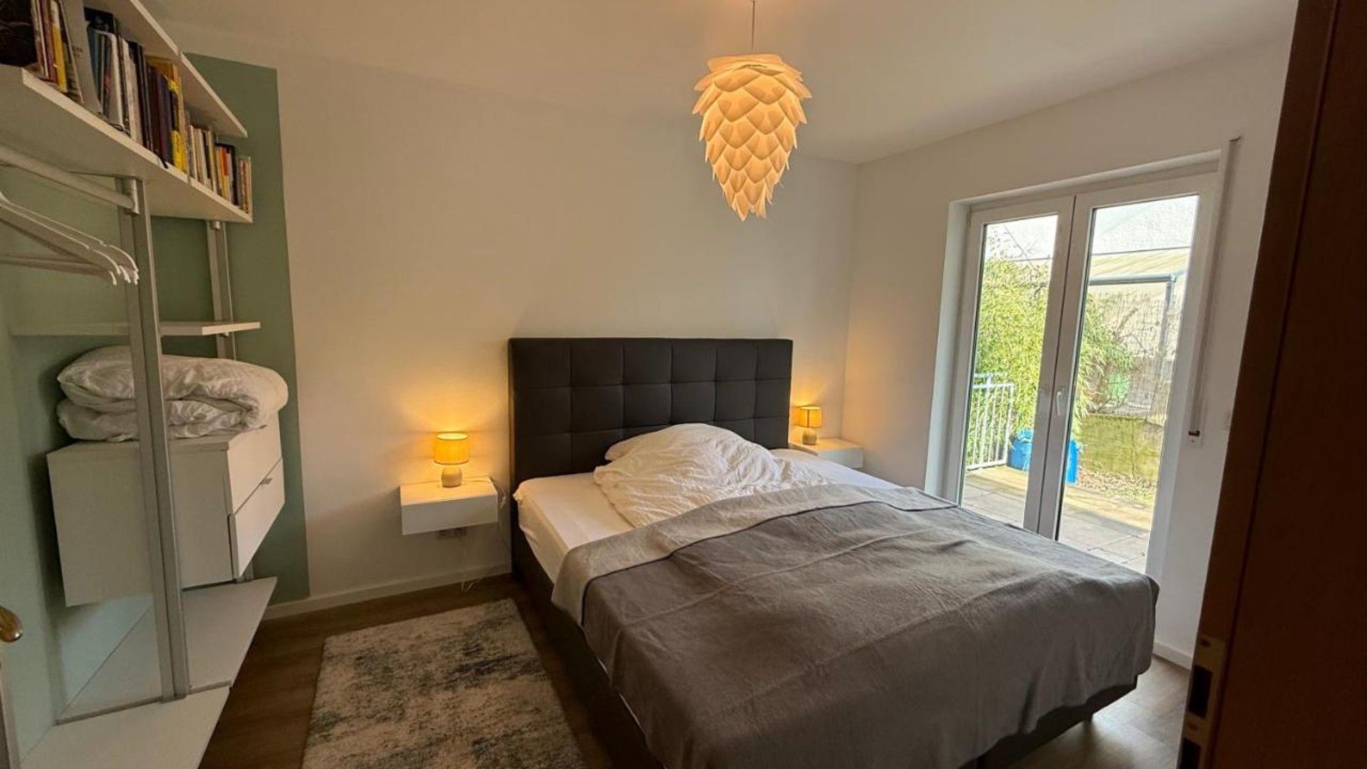Ein Schlafzimmer mit einem Bett, Nachttischlampen, einem Bücherregal und Blick durch eine Schiebetür.