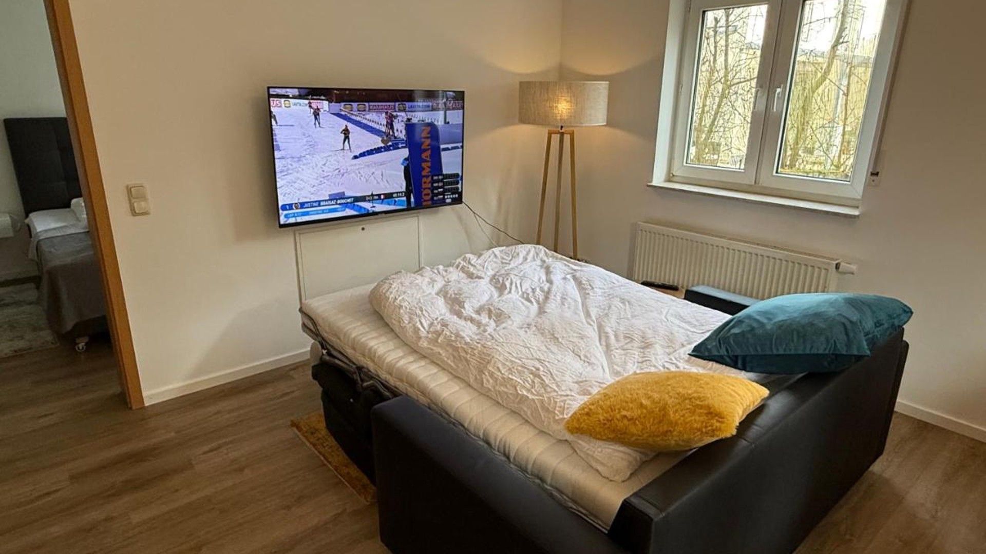 A bedroom with a bed, a television mounted on the wall, a window, and a floor lamp.