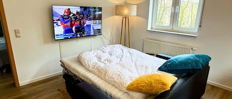 Un dormitorio con una cama, un televisor montado en la pared, una lámpara de pie y una ventana.