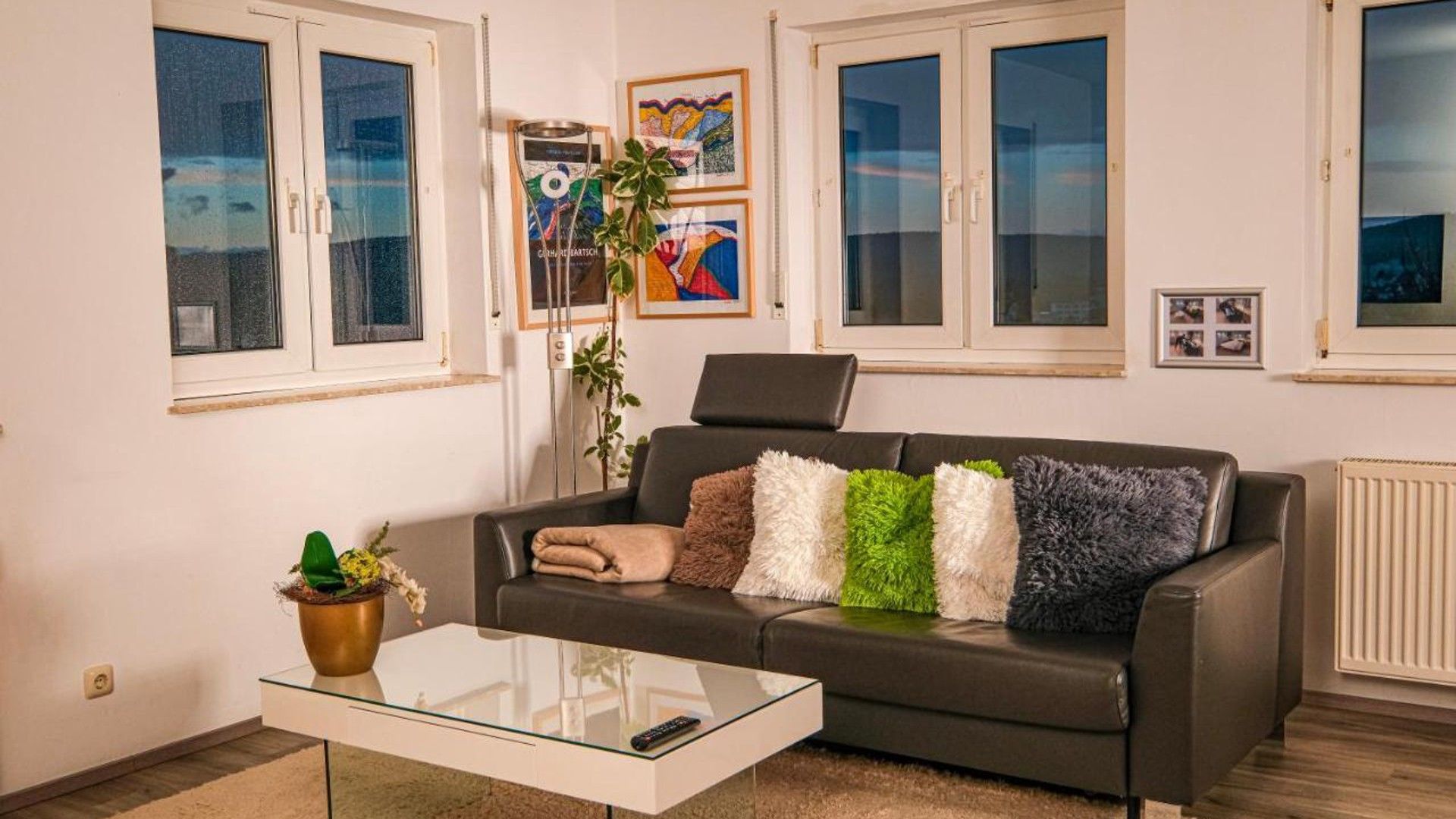 Un salon avec un canapé, une table basse en verre et une plante en pot. Il y a des fenêtres avec des rideaux et des œuvres d’art encadrées accrochées au mur.