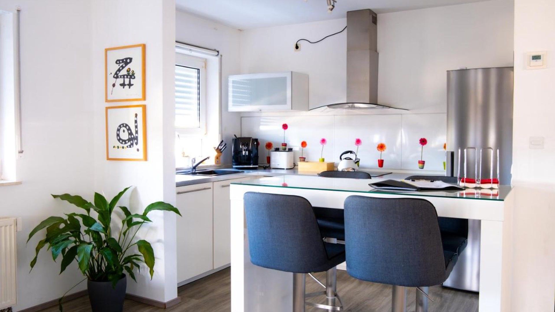 A modern kitchen with a central island, bar stools, a potted plant, and framed artwork on the wall.
