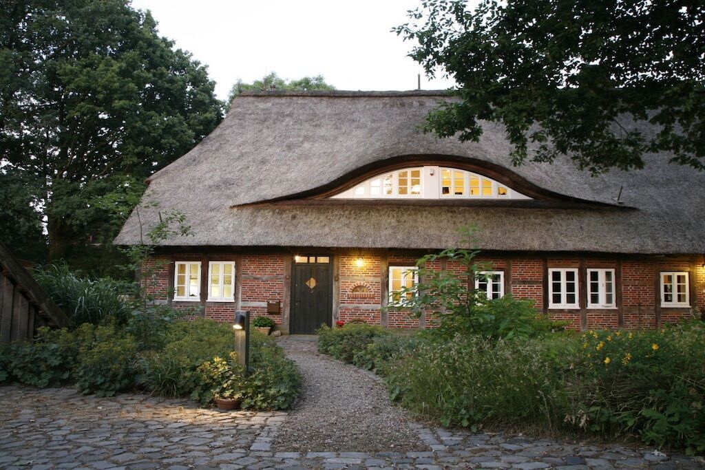 Une maison traditionnelle à toit de chaume, dotée d’une façade en briques, d’une porte centrale et d’un chemin menant à l’entrée.