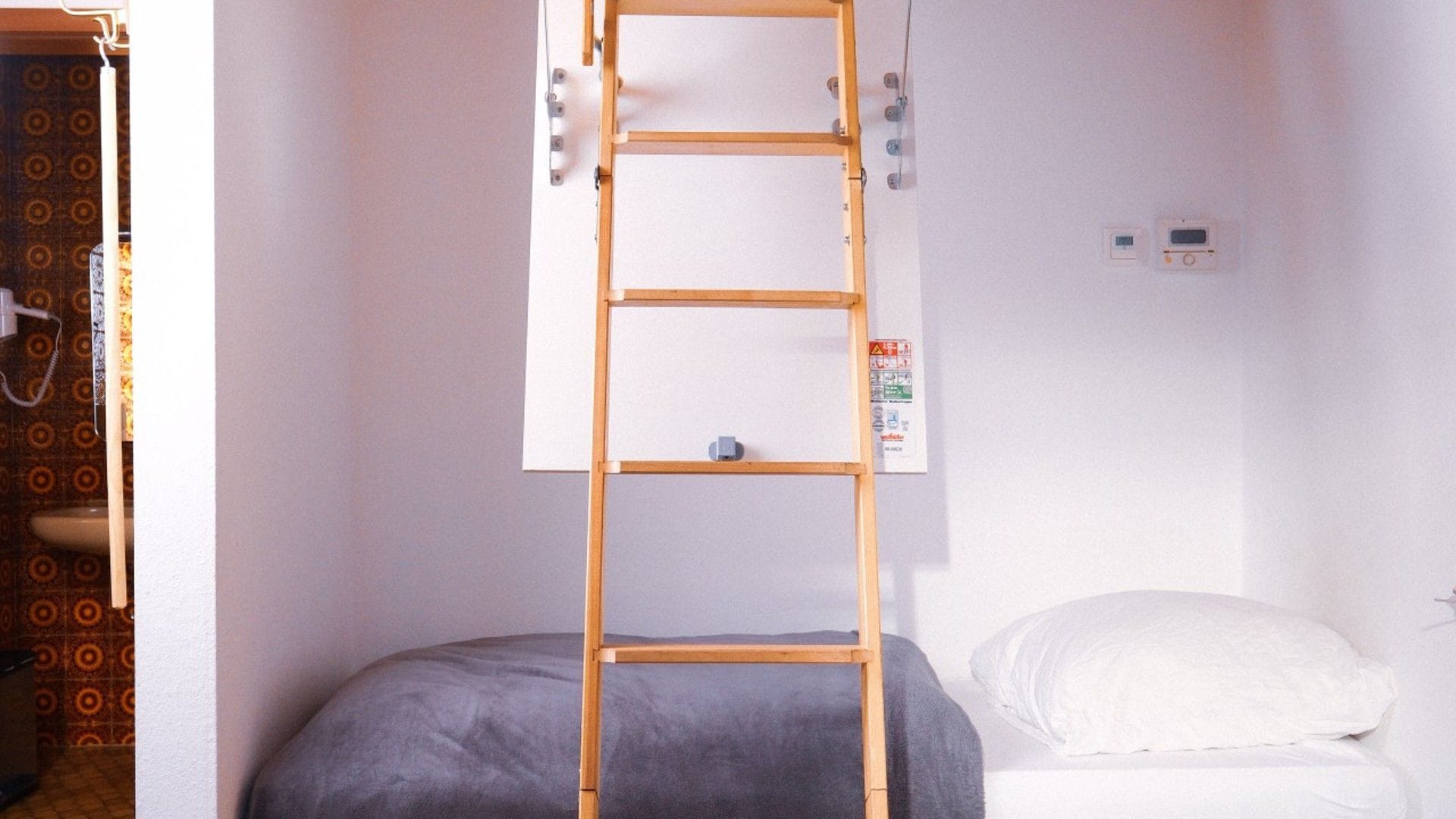 Ein Zimmer mit einer Holzleiter an der Wand, einem Bett mit einem weißen Kopfkissen und einem kleinen Wandthermostat.