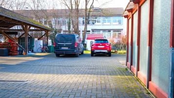 A paved driveway with two cars parked, a covered area with stacked items, and a building in the background.