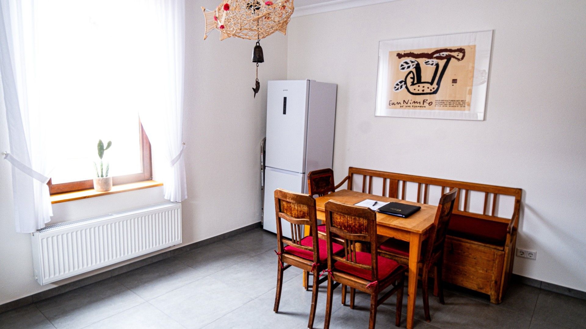 A dining area with a wooden table and chairs, a white refrigerator, and a window with a plant.