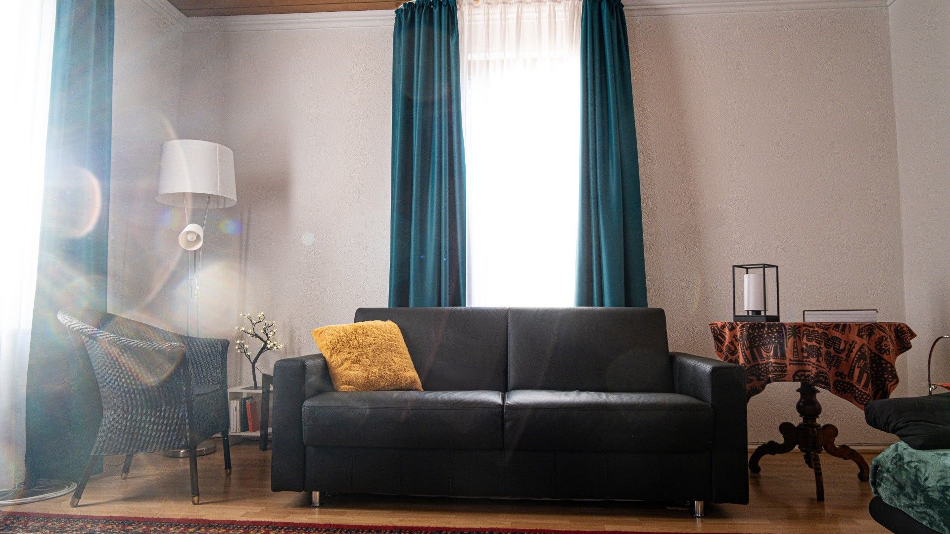 Ein Wohnzimmer mit einem schwarzen Sofa, einer Lampe, einem Tisch mit gemustertem Tischläufer und einem Sessel.