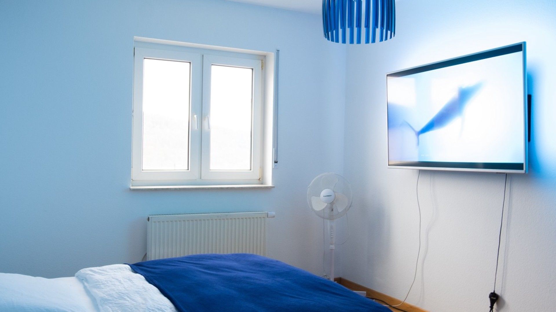 Une chambre avec un lit, une fenêtre, un téléviseur fixé au mur et un ventilateur sur pied.