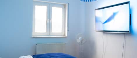 Ein Schlafzimmer mit Bett, Fenster, Wandfernseher und Standventilator.