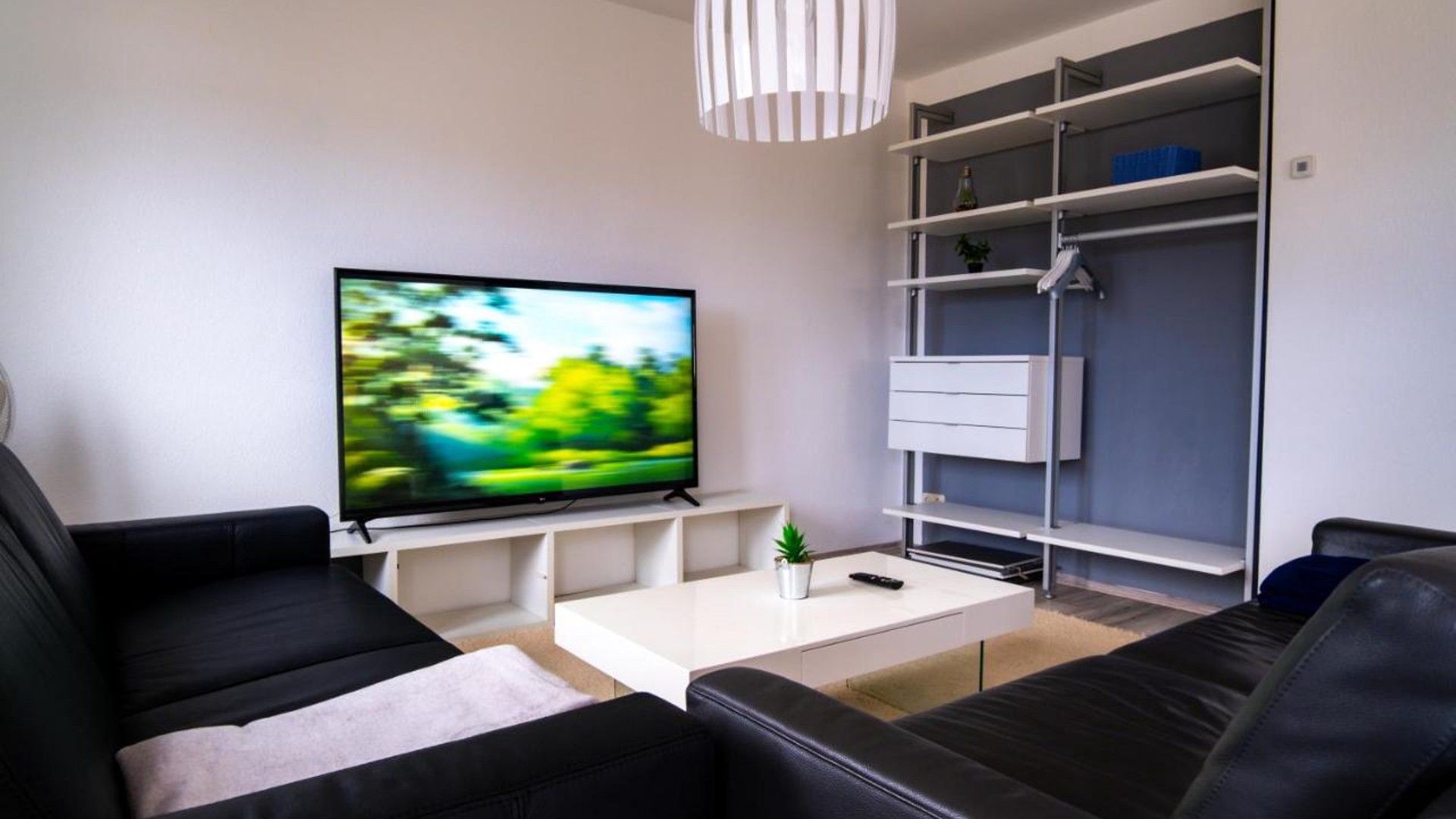 A modern living room with a flat-screen TV, black leather sofas, a white coffee table, and a grey shelving unit.
