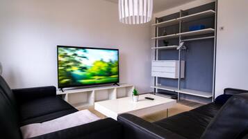 A modern living room with a flat-screen TV, black leather sofas, a white coffee table, and a grey shelving unit.