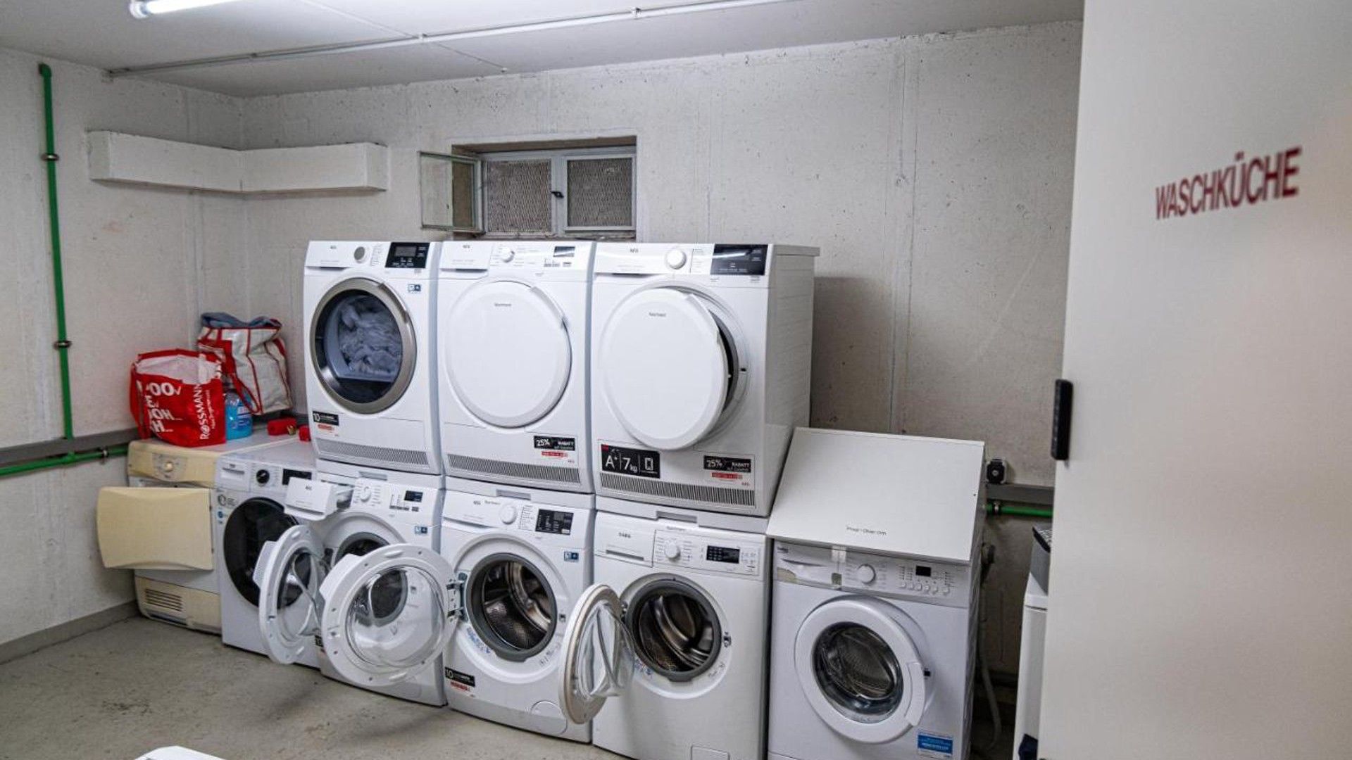 A laundry room with multiple washing machines and dryers, a ventilation system, and a red bag on the floor.