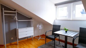 Ein Zimmer mit einem weißen Schreibtisch, zwei Korbstühlen, einem kleinen Tisch mit einer Pflanze, einem weißen Ventilator und einer weißen Regaleinheit.