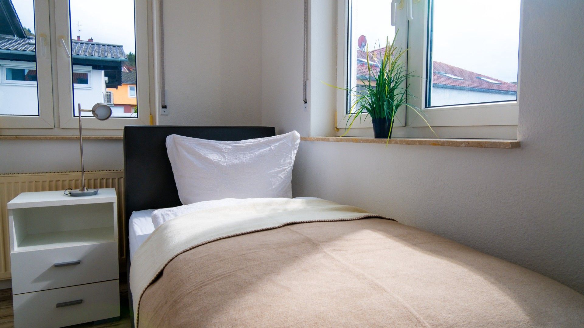 Ein Schlafzimmer mit Bett, Nachttisch, Lampe und einem Fenster mit Blick auf Gebäude.
