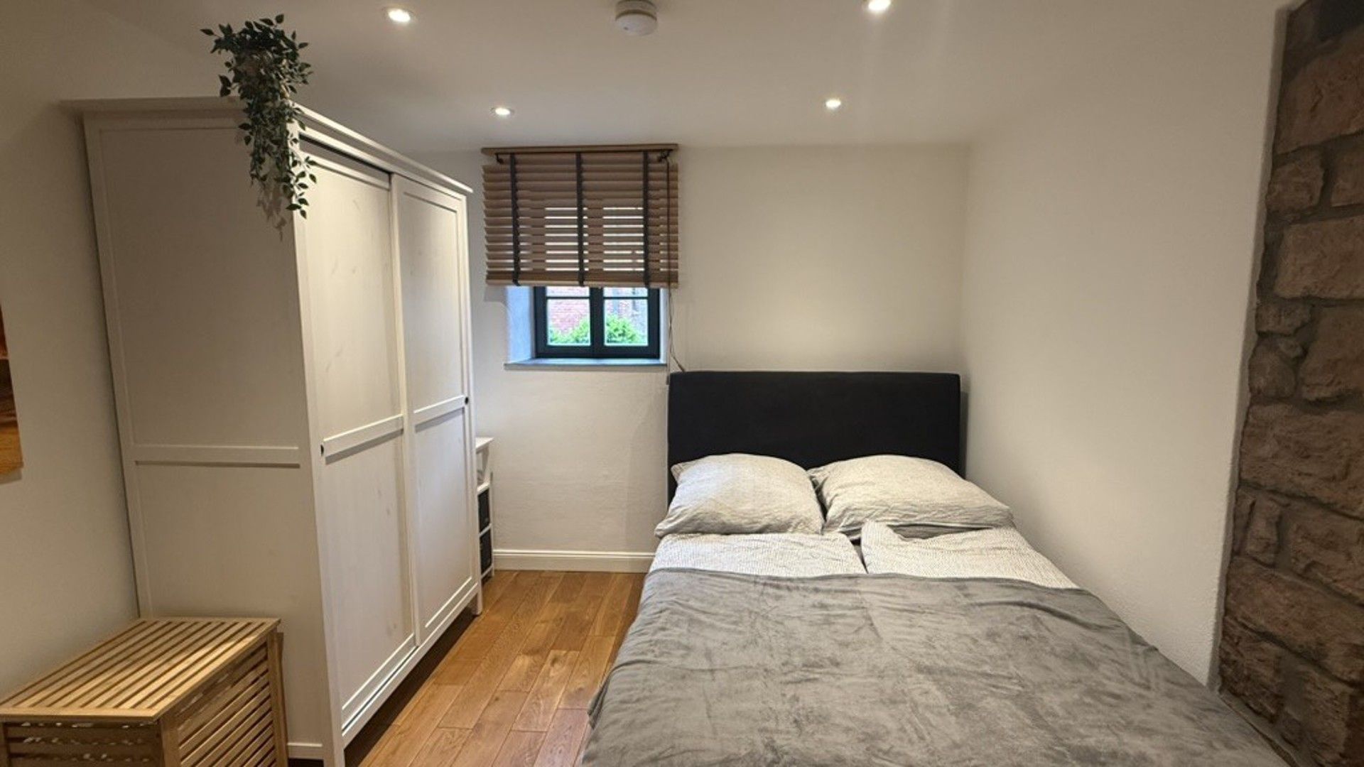Ein Schlafzimmer mit Bett, Fenster mit Jalousien, Kleiderschrank und einer kleinen Pflanze.