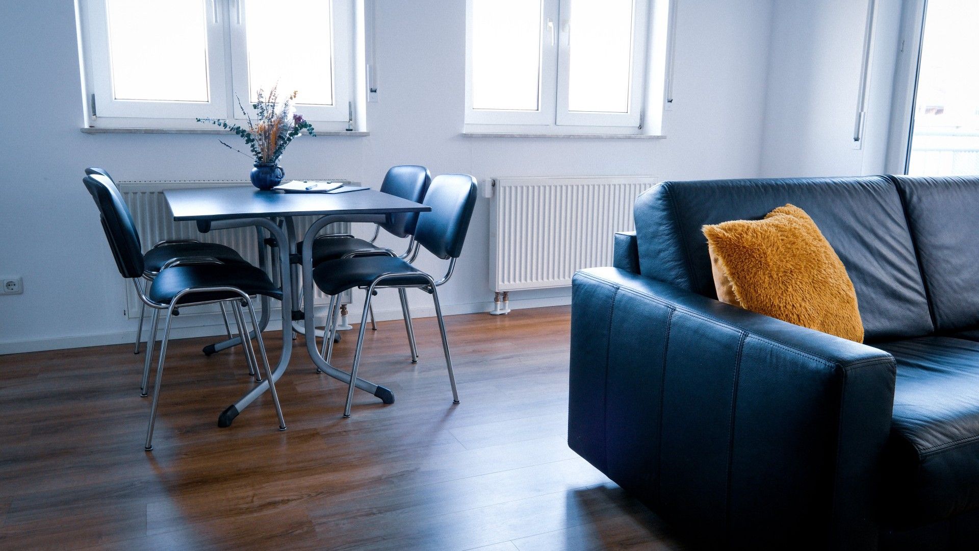 A modern living room with a blue leather sofa, a dining table with chairs, and a radiator.