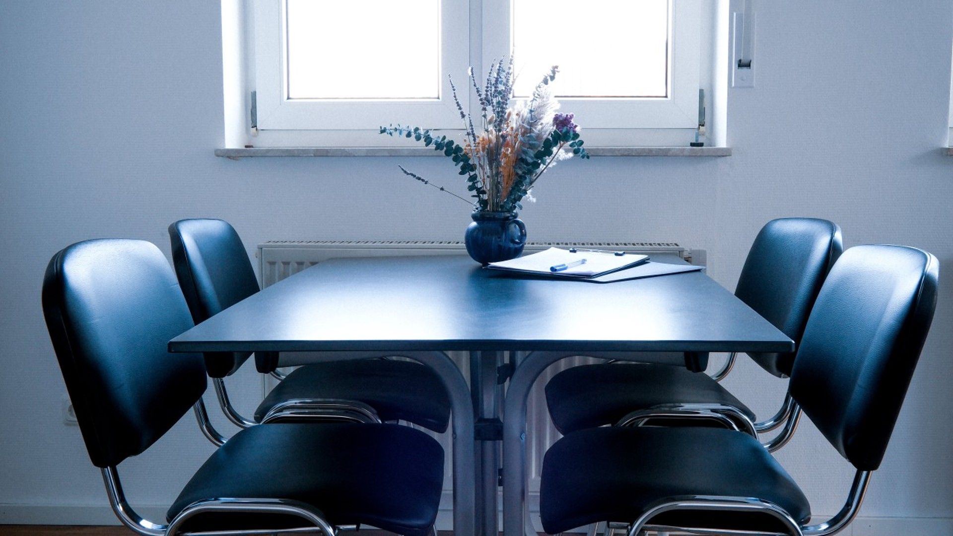 Uma sala de conferências com mesa, cadeiras, um vaso de flores e uma janela.