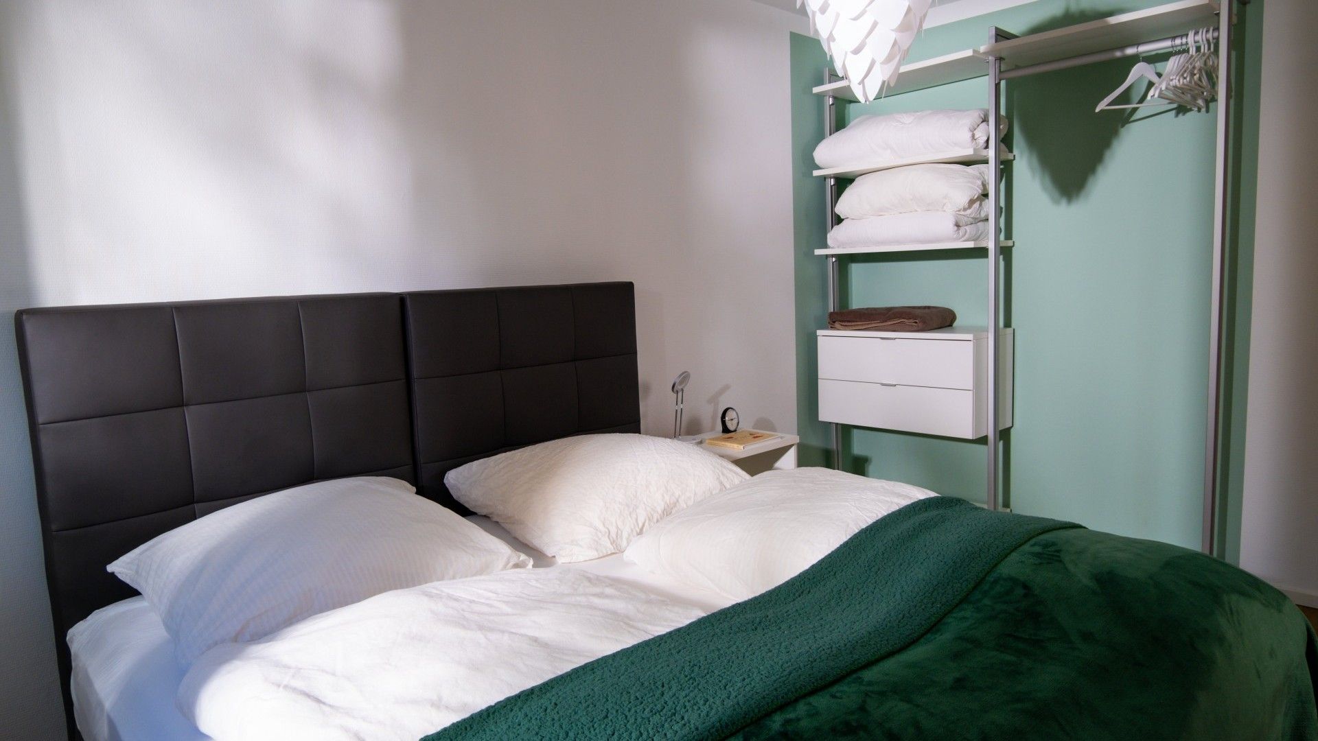 Ein Bett mit dunklem Kopfteil, weißer Bettwäsche und einer grünen Tagesdecke. Ein Nachttisch mit Lampe und Buch ist zu sehen. Ein weißes Regal zeigt gefaltete Handtücher und ein braunes Handtuch.