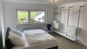 Quarto com cama, cadeira, guarda-roupa e janela com vista para uma casa e vegetação.