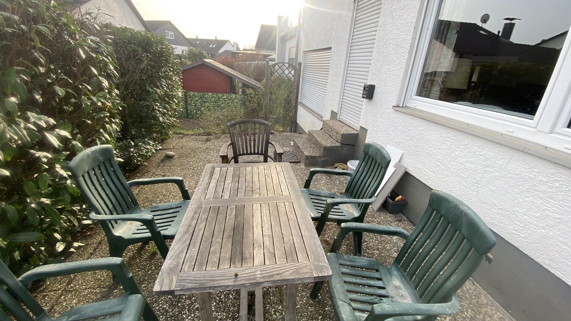 A patio with a wooden table and six chairs, surrounded by a hedge and a white building.