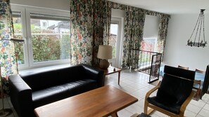 A living room with a black sofa, wooden table, and a chair. There are two windows with floral curtains, a lamp on a side table, and a hanging light fixture.