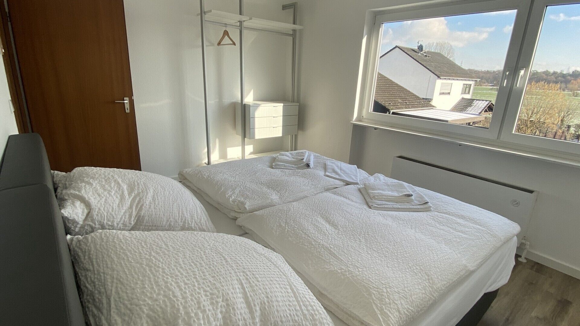 A bedroom with a bed, a bedside table, a hanging towel rack, and a window with a view of houses.