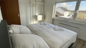 Un dormitorio con cama, mesita de noche, toallero y ventana con vistas a casas.