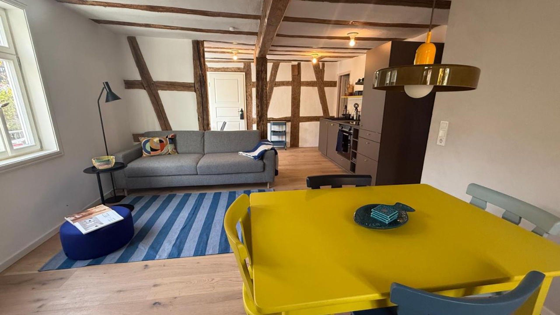 Ein Zimmer mit einem gelben Tisch, einem grauen Sofa, einem Streifen Teppich und freiliegenden Holzbalken an der Decke.