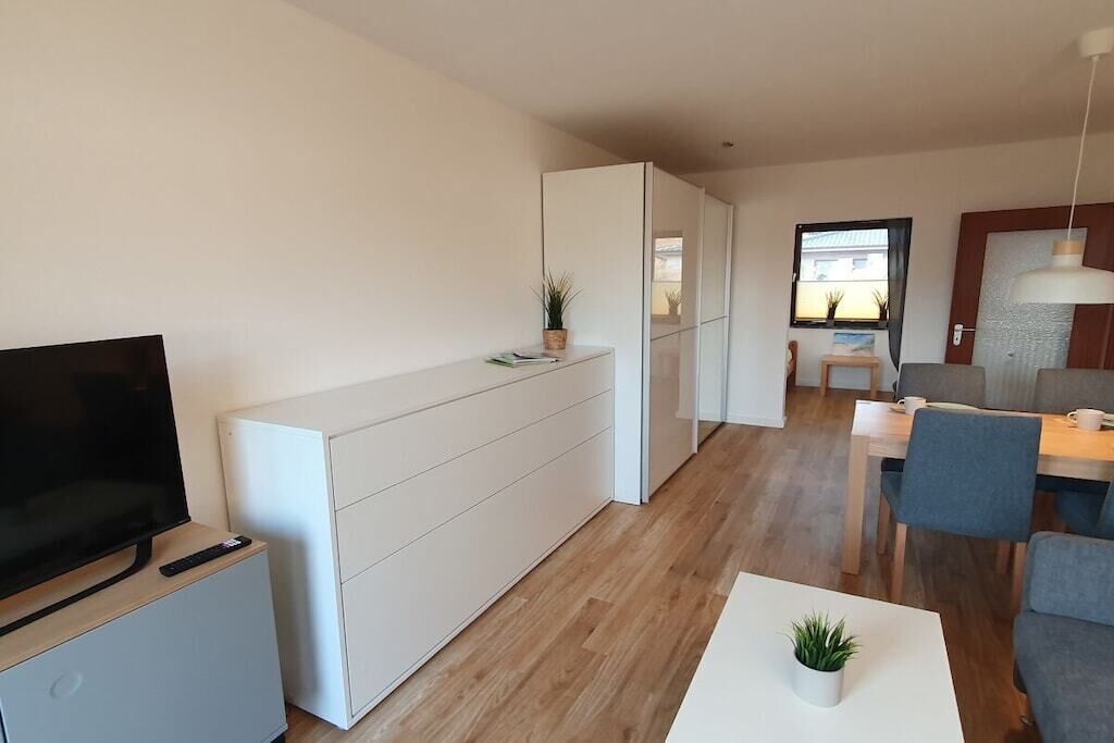 Una sala de estar moderna con televisor de pantalla plana, un mueble blanco, una zona de comedor con mesa y sillas, y una planta pequeña sobre la mesa de centro.