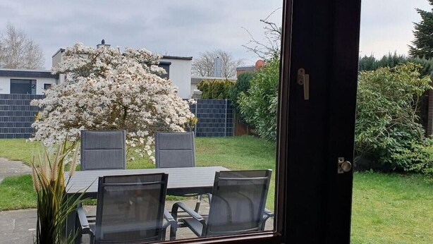 Blick durch eine Glastür auf einen Patio mit Tisch und Stühlen, einen blühenden Baum und einen Hecken.