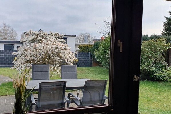 On aperçoit, à travers une porte vitrée, un patio avec une table et des chaises, un arbre en fleurs et une haie.