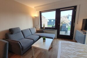 A modern living room with a grey sofa, a white coffee table, and a bed. There is a large window with a view of the outside. - Holiday apartment with 2 bedrooms (Cuxhaven)