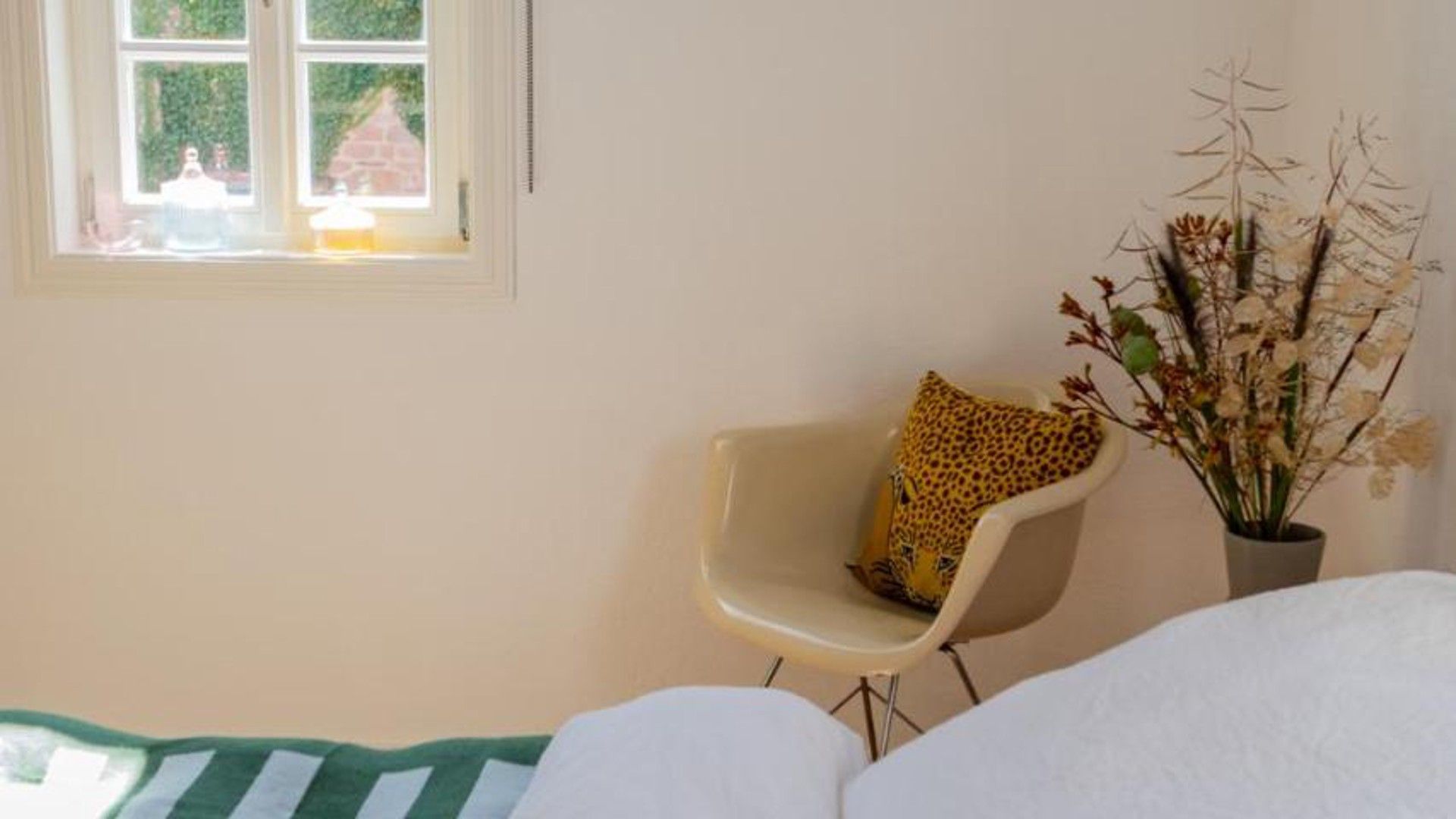 Ein Zimmer mit einem Bett, einem Sessel mit Leopardenmuster-Kissen, einem Fenster mit Blick auf Grünflächen und einer Topfpflanze.
