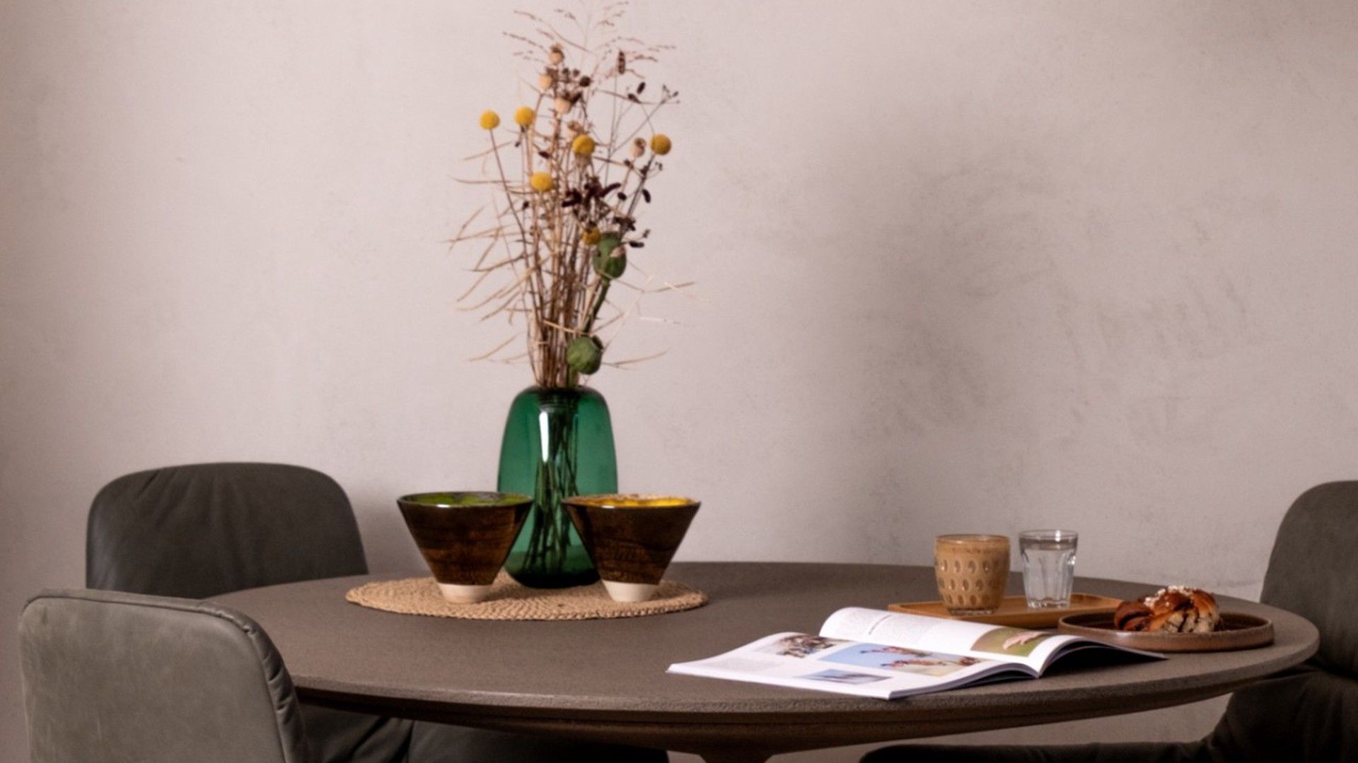 A round table with a magazine, a bowl, a glass, and a decorative vase with dried plants.