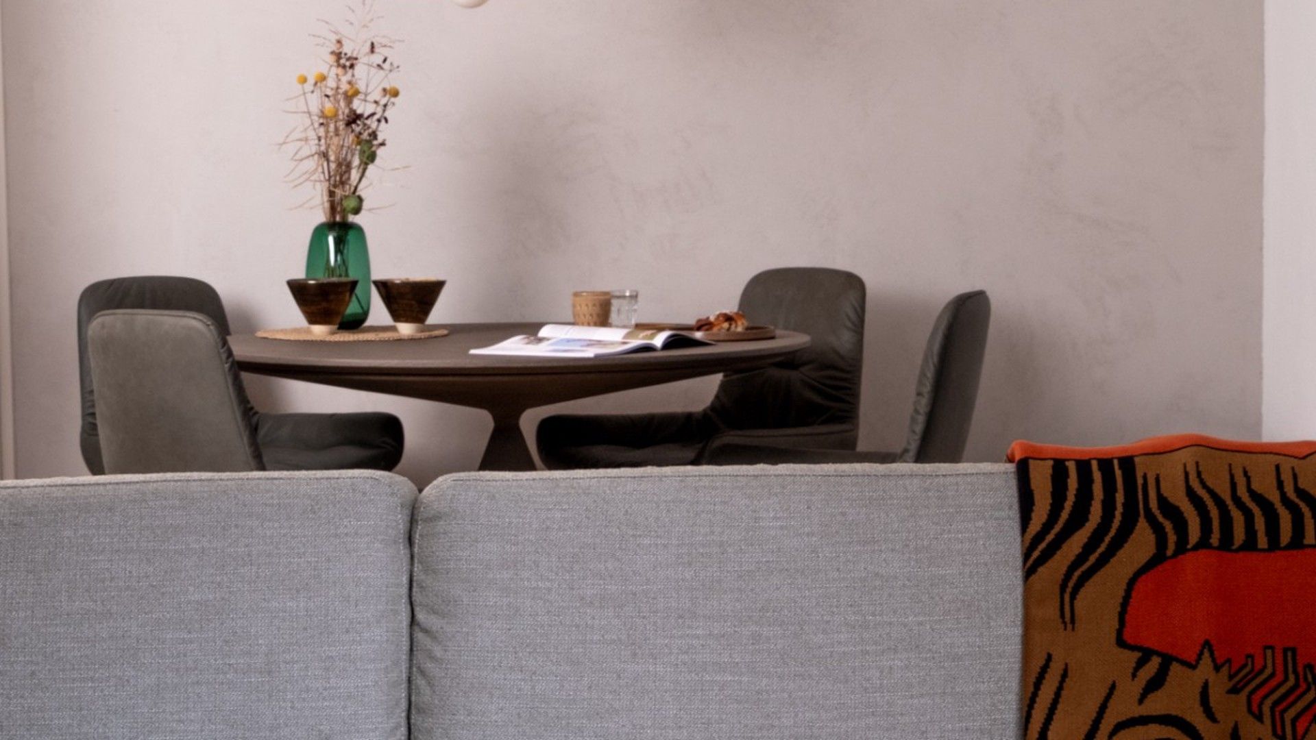 A dining area with a round table, two chairs, a vase with dried flowers, and a book.