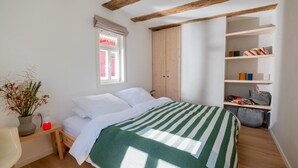 Een slaapkamer met een bed, een raam, een kledingkast en een plank met tassen.