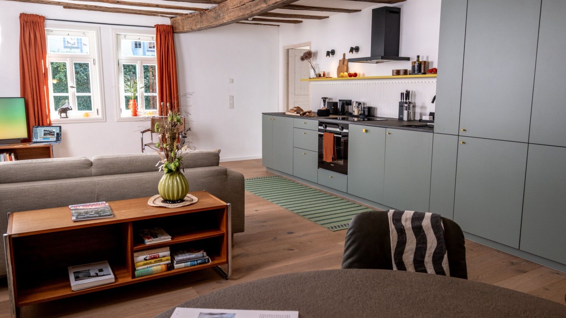 A modern living room with a sofa, a coffee table with books, a green vase, and a kitchen with grey cabinets and a black range hood.