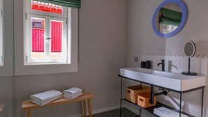 Een badkamer met een witte wastafel, een spiegel, een raam met rode gordijnen en een houten bankje met handdoeken.