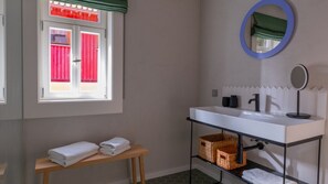 Ein Badezimmer mit weißem Waschbecken, einem Spiegel, einem Fenster mit roten Vorhängen und einer hölzernen Bank mit Handtüchern.