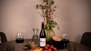 Una mesa de comedor con una botella de vino, copas, un florero con plantas secas, un cuenco de uvas y una cesta de pan.