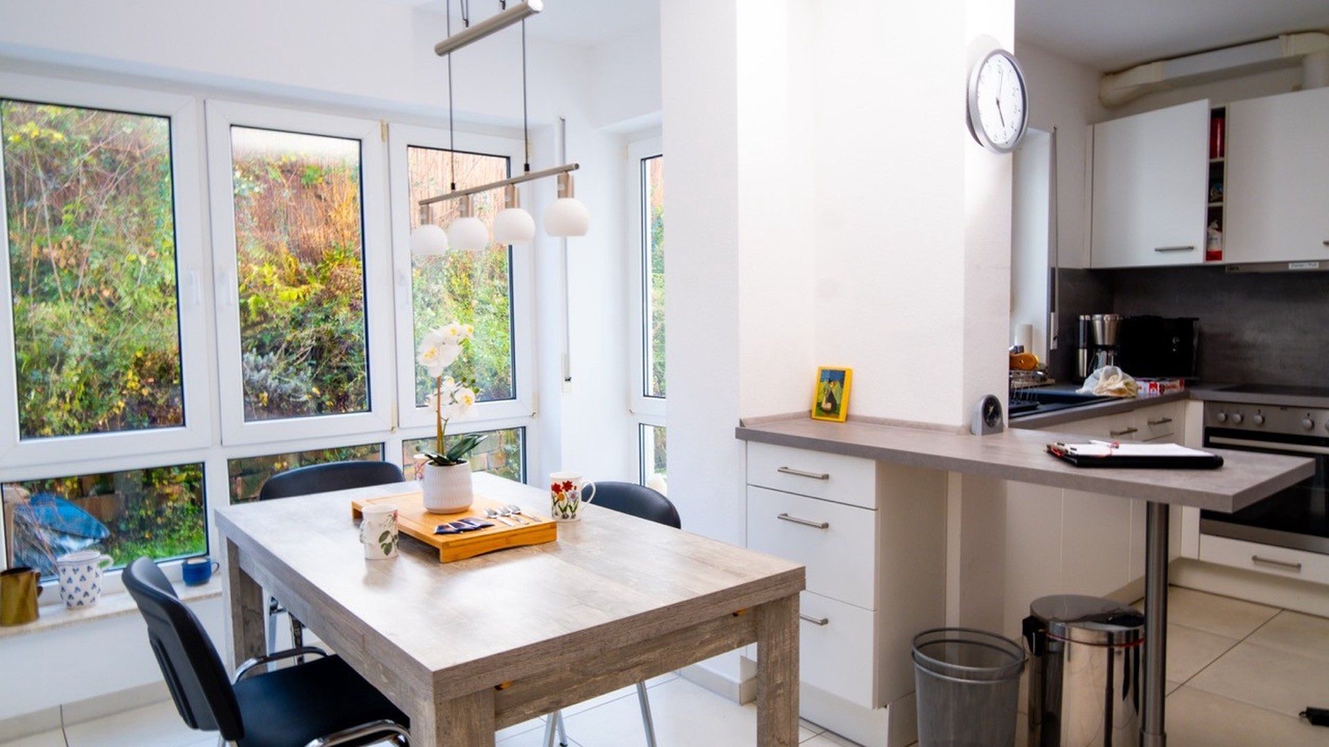 A modern kitchen with white cabinets, a wooden dining table, and a view of greenery through the windows.