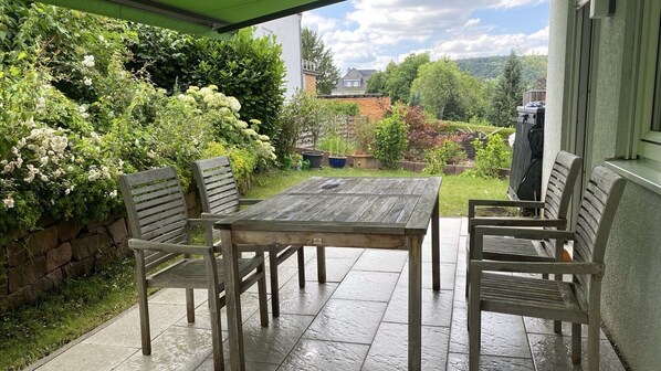 Un patio al aire libre con una mesa y sillas de madera, rodeado de exuberante follaje y un muro de piedra.
