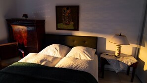 Un dormitorio con cama, mesita de noche con lámpara, cómoda y un cuadro en la pared.