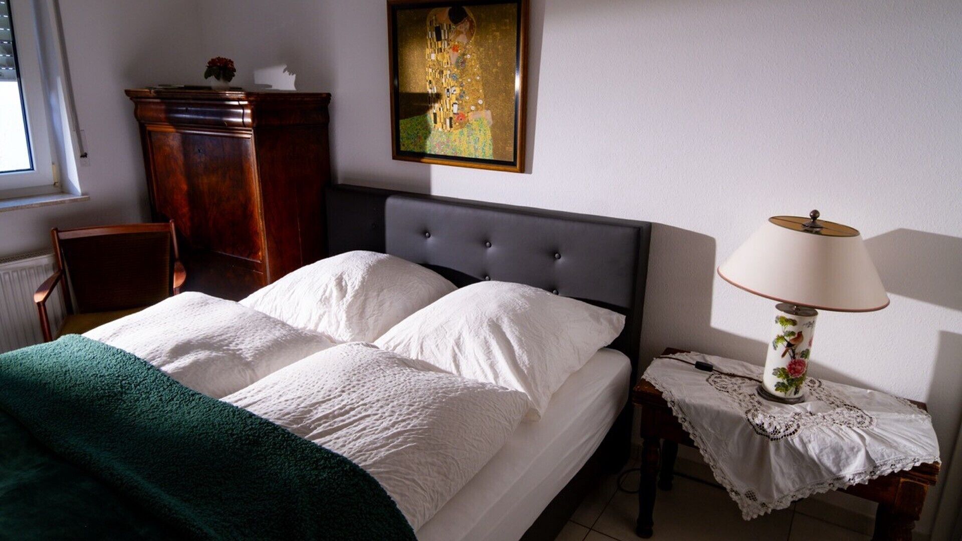 Ein Schlafzimmer mit einem Bett, einem Nachttisch mit Lampe, einem Sessel, einer Kommode und einem Bild an der Wand.