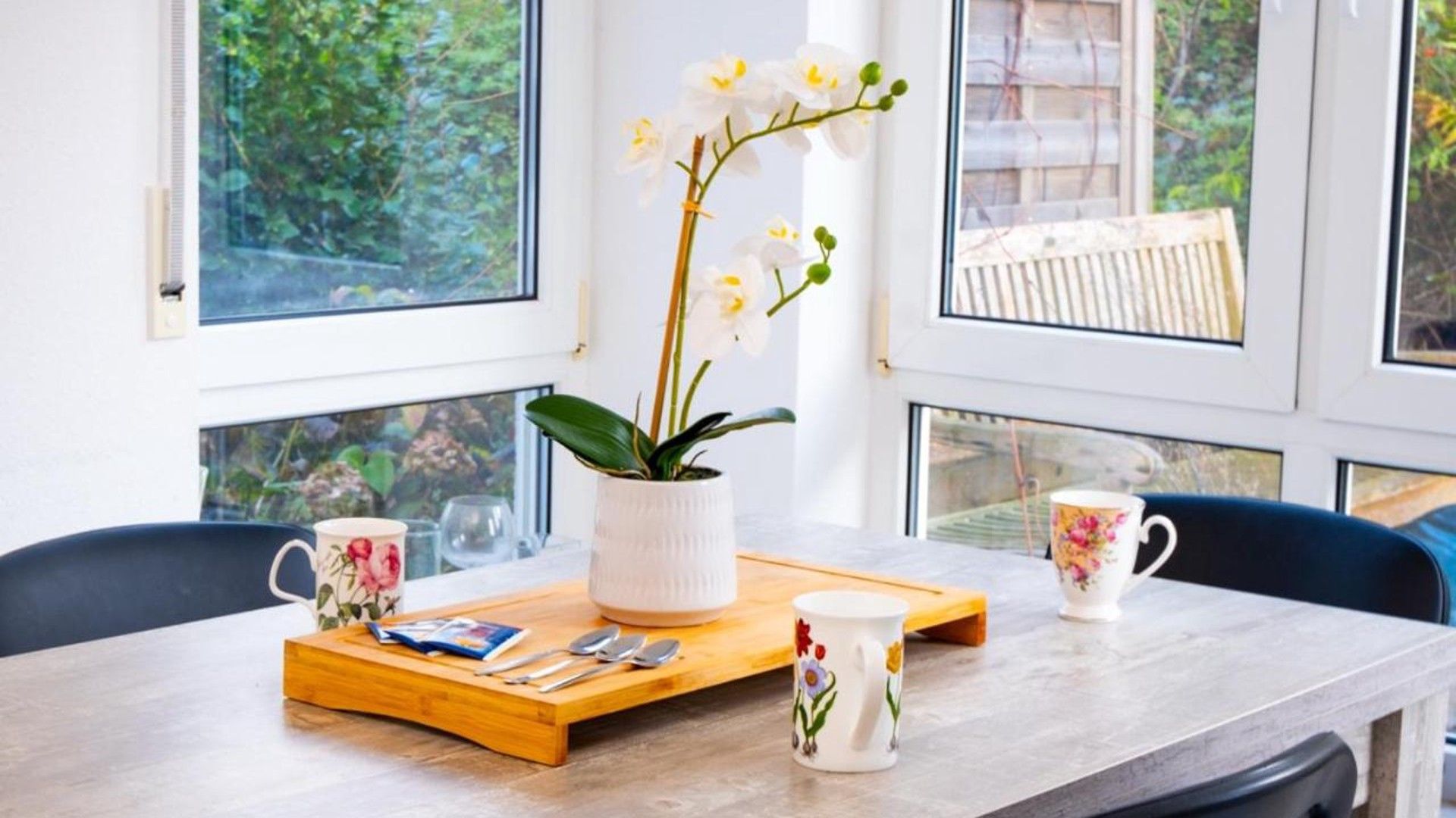 A dining table with a white vase of orchids, two floral mugs, and a wooden tray with cutlery.