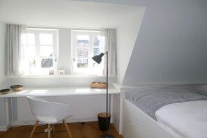 Une chambre à coucher avec un lit, un bureau avec une chaise, une fenêtre avec des rideaux et une lampe sur pied.