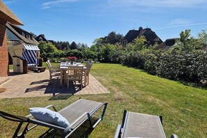 Une terrasse avec des meubles d’extérieur, une table décorée de fleurs et un espace gazonné avec des chaises longues.