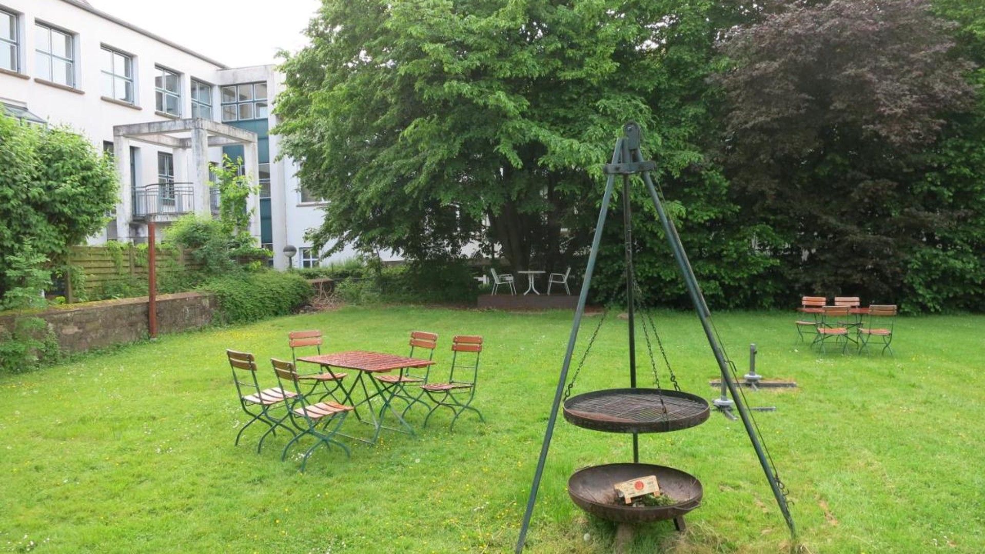 Ein grünes Freigelände mit einer Schaukel, einem runden Tisch mit Stühlen und einer kleinen Feuerstelle.