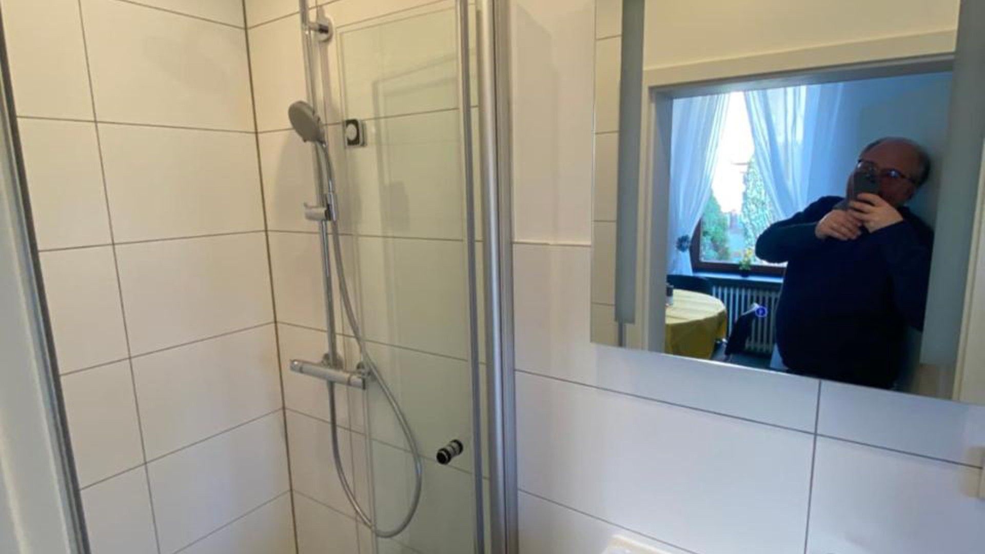 Ein Badezimmer mit Duschkabine aus Glas, einem Händetrockner und einem Spiegel, der eine Person reflektiert.